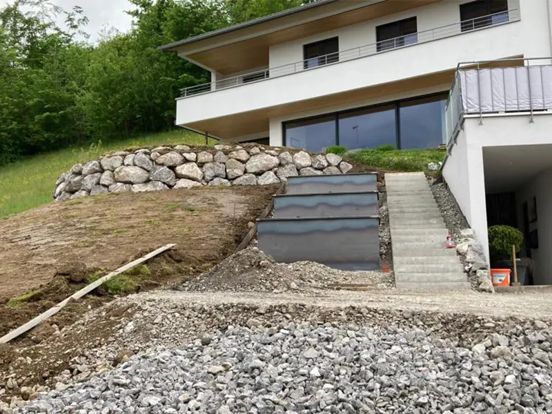 Hangbefestigung mit Naturstein-Stützmauer, Treppenanlage und vorbereiteten Erdflächen vor einem Wohnhaus.