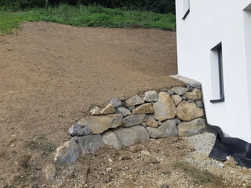 Teilfertige Natursteinmauer zur Hangsicherung neben einem Wohngebäude.