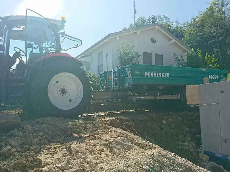 Traktor mit Anhänger beim Materialtransport auf einer Baustelle neben einem Wohnhaus.