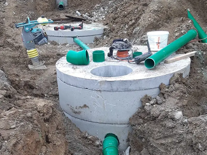 Einbau eines Kontrollschachts mit angeschlossenen grünen Abwasserrohren in offener Baugrube.