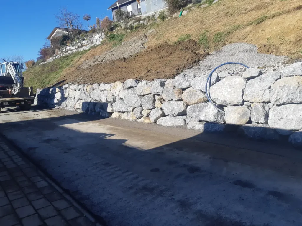 Naturstein-Stützmauer entlang einer Straße zur Hangsicherung mit frisch modelliertem Erdreich oberhalb.