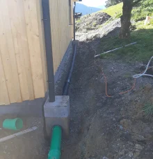 Drainageleitung entlang eines Holzgebäudes mit Anschluss an ein grünes Abwasserrohr.